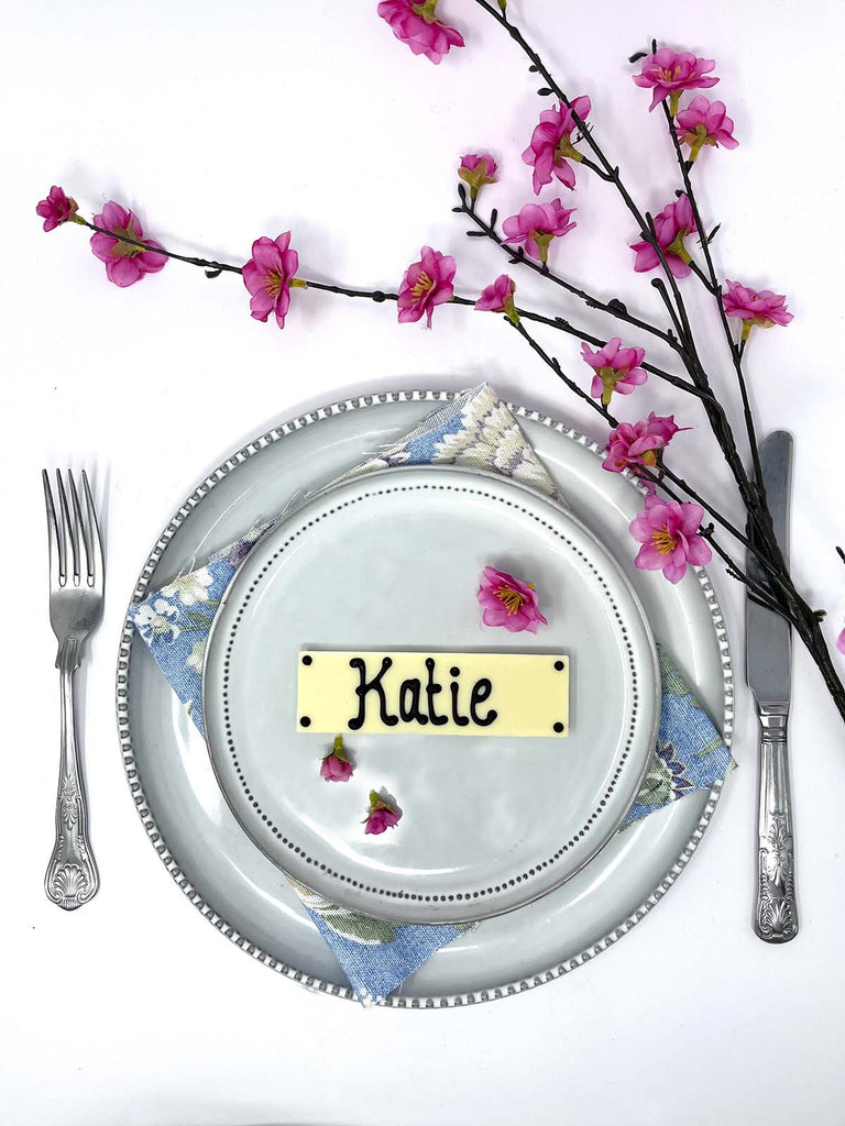Decorative place setting with a white chocolate bar as a name card labeled 'Henry', silverware, and cherry blossom branches on a white background.
