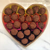 A heart-shaped box containing multiple dark chocolate truffles with cocoa powder dusted on top, displayed on a marble surface.